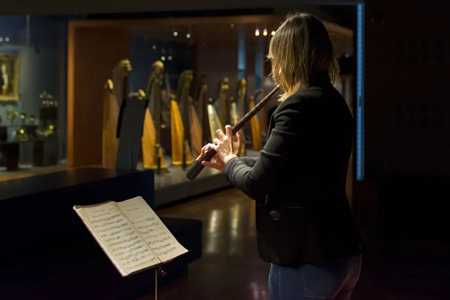Eva Godard jouant du cornet dans les salles du Musée de la musique, devant des harpes anciennes